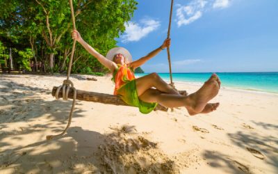 Happy and attractive woman with colorful sarongs and white wide-brimmed hat swinging on tropical white beach of Koh Rok Islands, Ko Lanta, Thailand paradise for snorkelers and divers.